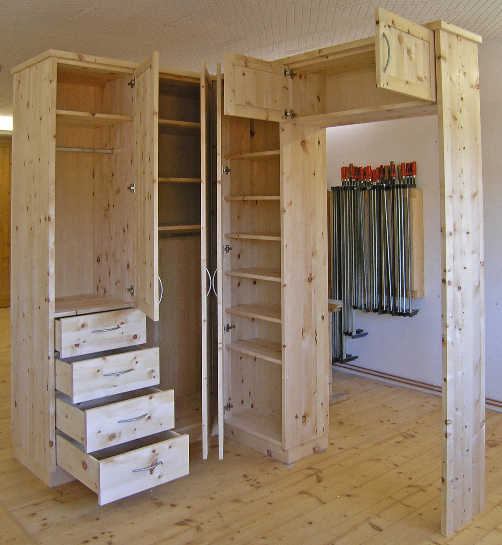 Blick in den Zirbenholz-Kleider-Eckschrank 09.8, aus massiver, hochwertigen Tiroler Alpenzirbe. 3 Türen, 4 Schubladen plus 2 Fächer über der Zimmertüre, Fronten mit abgeplatteten Füllungen und gebogenen Messingriffen, Sockel, Maßeinbau auf Deckenhöhe, Türüberbau